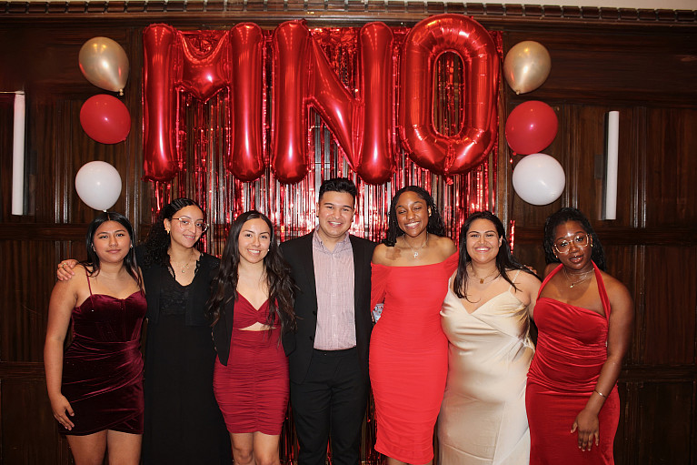 A group of students from different backgrounds in formal attire at a celebration with red balloons spelling MNO